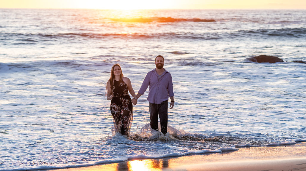 Engaged couple holding hands walking away from sunset and waves.