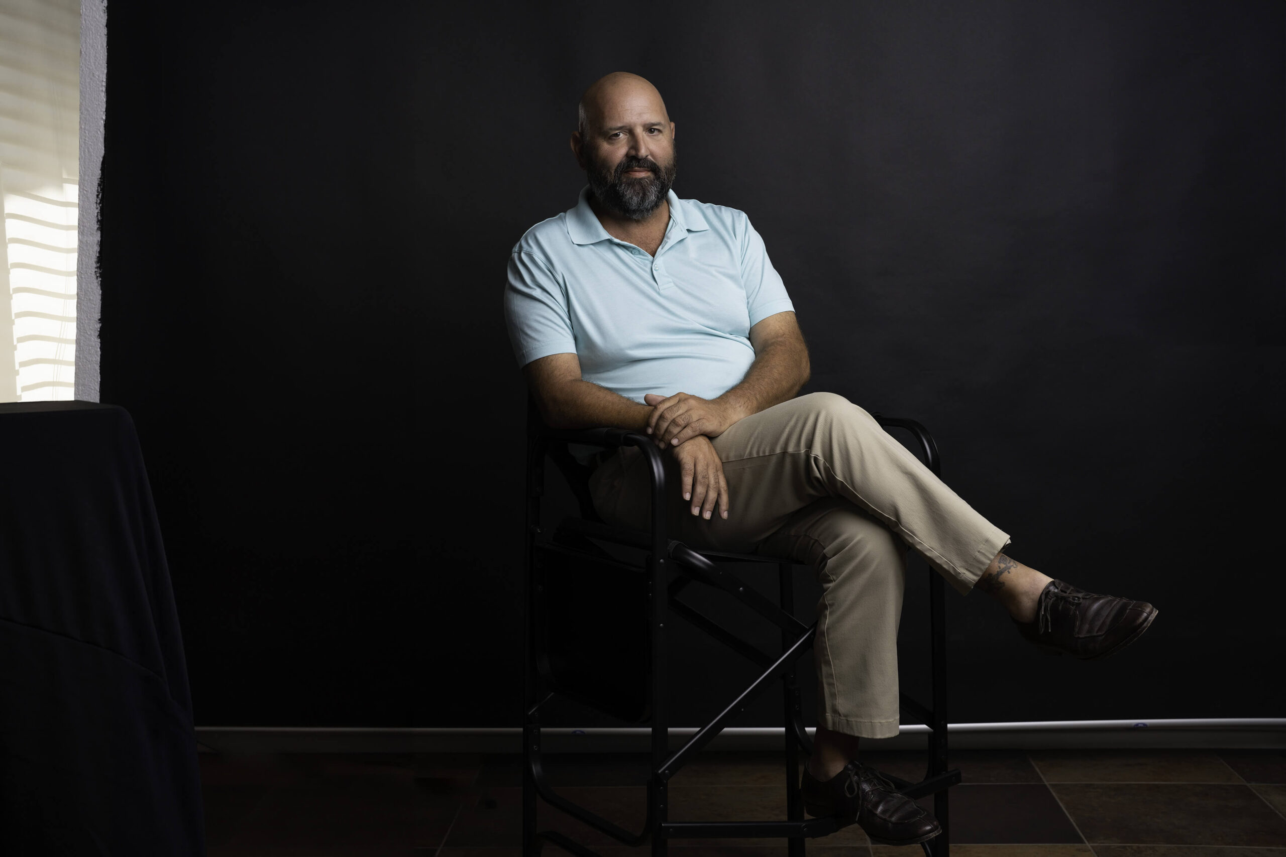 "A confident professional man sits in a director's chair against a dark, seamless background in a Riverside headshot studio. His attire, a light blue polo shirt and khaki pants paired with dark brown shoes, suggests a casual business style suitable for an approachable Riverside headshot.