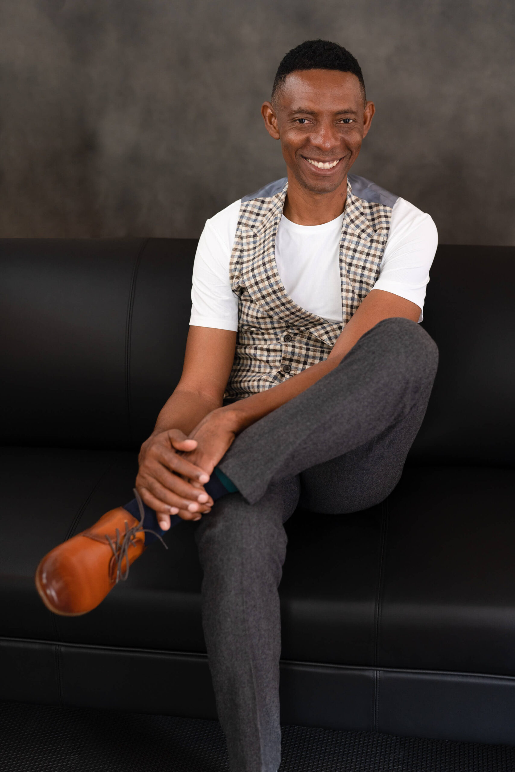 Smiling man getting a loma linda headshot, in fashionable vest and grey trousers seated on a black couch for a relaxed yet professional branding portrait, in a Loma Linda photography studio.